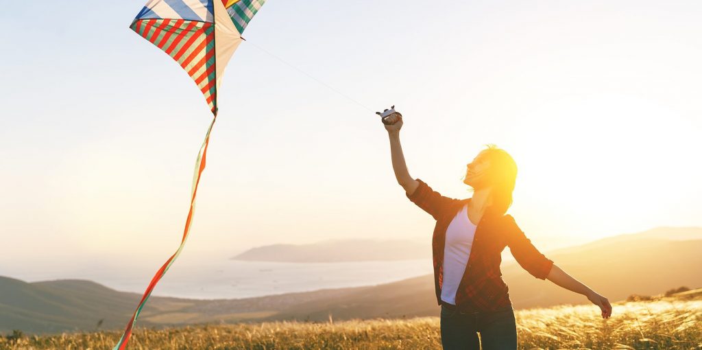 Happy young woman running with a kite on a glade at sunset in summer